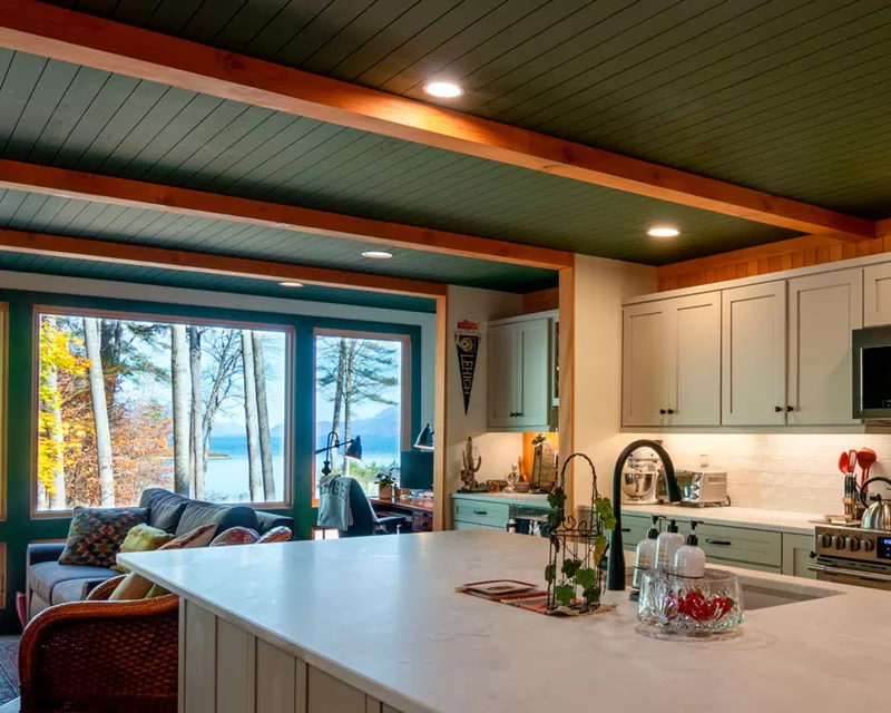 A cozy kitchen and living space with a lake view through large windows. The kitchen features a white marble island with a black faucet, soap dispensers, and a decorative plant. The cabinetry is light sage green with black knobs, and the backsplash consists of white subway tiles. The ceiling is painted dark green with exposed wooden beams. The living area includes a sofa with colorful pillows, a wicker chair, and a desk with a lamp near the window. The background showcases tall trees and a lake with mountains in the distance.