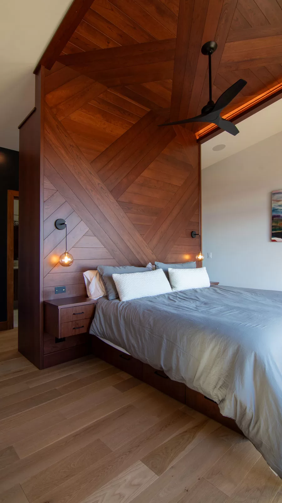 Modern master bedroom in Cinderwave, designed by AJA Architecture and Planning. This striking space features a custom wood accent wall with geometric paneling that extends seamlessly into the vaulted ceiling, creating a cohesive and warm aesthetic. Integrated floating nightstands enhance the sleek, built-in design, while pendant lighting with brass and glass elements adds a soft, ambient glow. A matte black ceiling fan contrasts beautifully with the rich wood tones, reinforcing the modern yet natural aesthetic. Soft gray bedding and minimalist décor keep the space serene and inviting. The thoughtful combination of craftsmanship, texture, and lighting makes this bedroom a luxurious and tranquil retreat.