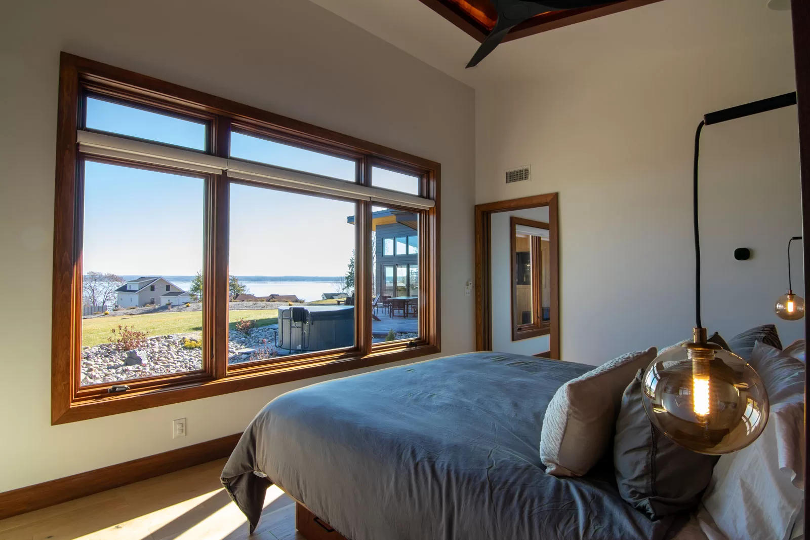 Luxury bedroom with lake views in Cinderwave, designed by AJA Architecture and Planning. This serene retreat features a modern, minimalist design with a focus on natural light and scenic vistas. Floor-to-ceiling windows framed in rich wood provide breathtaking views of the water and surrounding landscape. A contemporary bed with soft gray linens and plush pillows enhances the cozy yet sophisticated atmosphere. Pendant lighting with brass and glass elements adds warmth and elegance. The space seamlessly connects to an outdoor patio with a hot tub, creating a perfect indoor-outdoor living experience. This bedroom embodies tranquility, modern design, and a strong connection to nature.