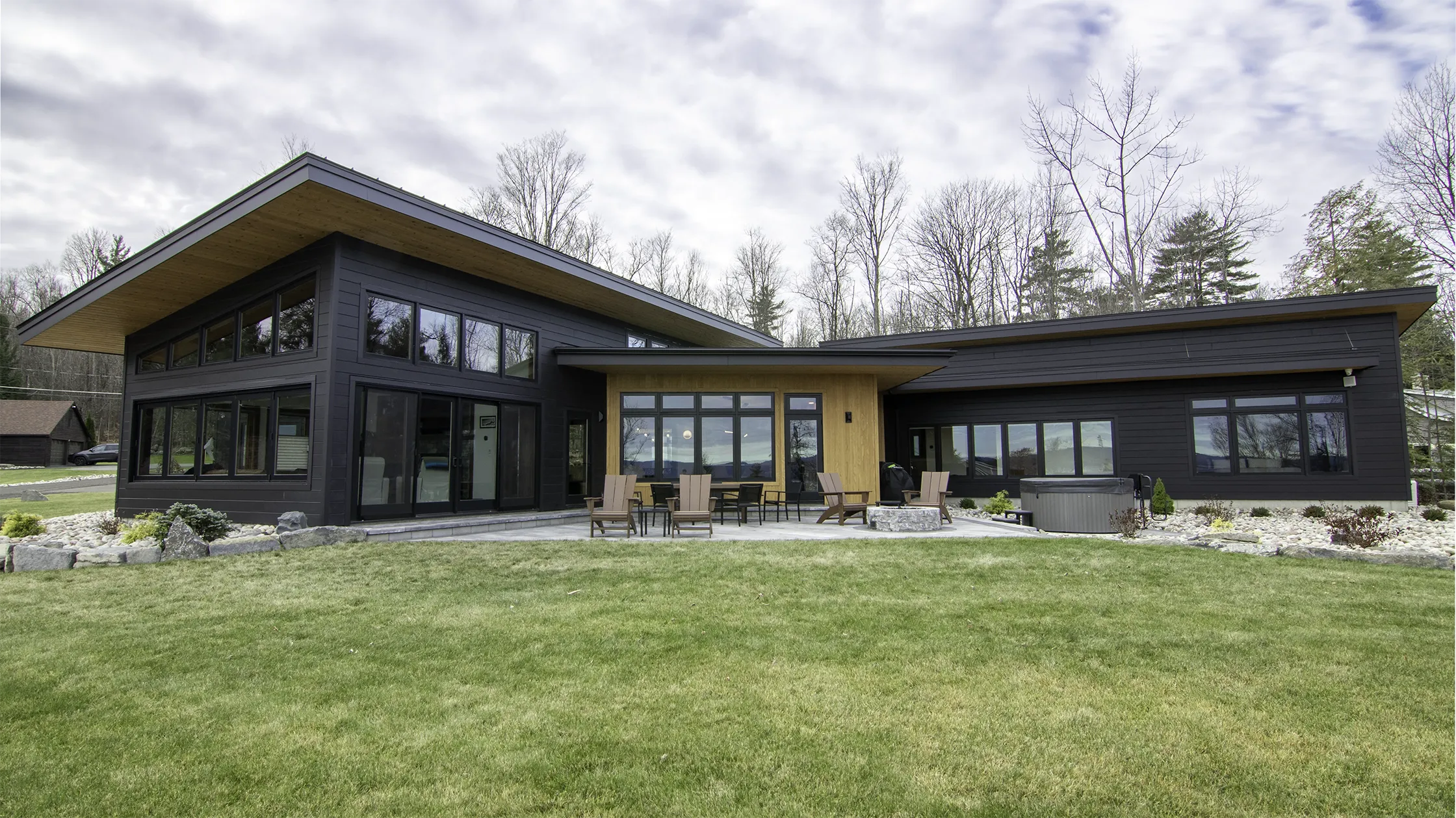 Cinderwave by AJA Architecture and Planning is a modern lakefront home with a sleek black exterior and natural wood accents, featuring large windows and sloped rooflines. The outdoor patio area includes Adirondack chairs and a fire pit, with a hot tub positioned to the right. The home is surrounded by a well-manicured lawn, landscaping with stone accents, and a backdrop of leafless trees under a cloudy sky.