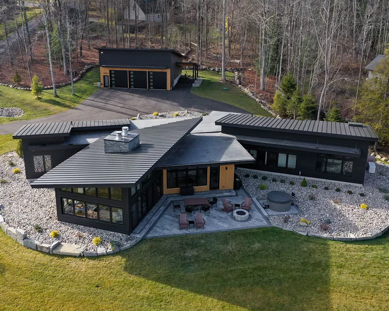 Modern lakefront home named "Cinderwave," designed by AJA Architecture and Planning. The residence features a striking black metal roof with angular forms, dark exterior cladding accented with natural wood, and expansive glass windows. A stone-lined landscape surrounds the home, complementing the sleek design. A spacious patio extends from the rear, equipped with outdoor seating, a fire pit, and a hot tub. A detached three-car garage sits at the top of the driveway, blending seamlessly with the modern aesthetic. The home is nestled among a wooded backdrop, offering a secluded and serene atmosphere.