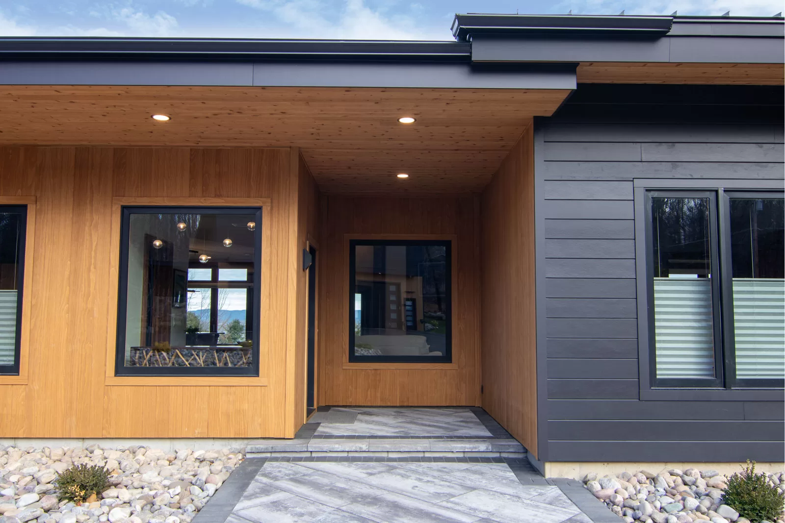 Modern home entryway in Cinderwave, designed by AJA Architecture and Planning. This striking façade features a dynamic contrast between warm vertical wood siding and sleek black horizontal paneling, creating a contemporary yet inviting aesthetic. The recessed entrance is framed by a wood-paneled soffit with integrated lighting, enhancing the welcoming atmosphere. Large black-framed windows provide glimpses of the home's interior while allowing natural light to filter through. The entrance path is composed of bold, textured stone pavers with dark accent borders, seamlessly integrating with the surrounding landscape. This modern entryway blends organic materials with refined architectural details for a sophisticated and timeless curb appeal.