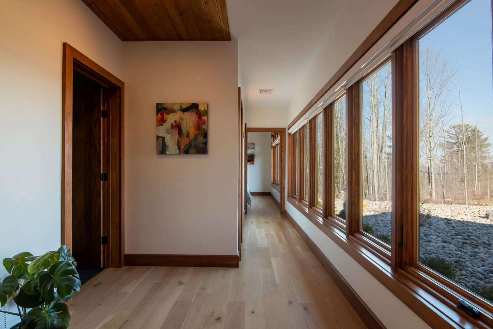 Modern hallway design in Cinderwave, created by AJA Architecture and Planning. This light-filled corridor features expansive floor-to-ceiling windows framed in rich wood, offering a seamless connection to the natural landscape outside. The warm oak flooring and wood-paneled ceiling add depth and warmth to the contemporary design. A minimalist approach to decor includes an abstract painting and a lush green plant, enhancing the organic and modern aesthetic. This thoughtfully designed hallway balances natural materials, abundant natural light, and architectural elegance, creating a tranquil and inviting passageway within the home.