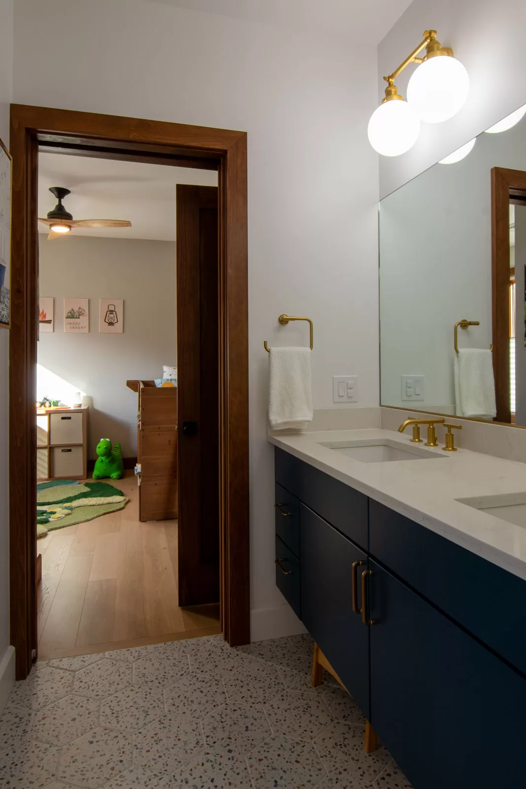 Modern kids' bathroom in Cinderwave, designed by AJA Architecture and Planning. This playful yet sophisticated space features a navy blue floating vanity with gold hardware, a white quartz countertop, and a double sink for added functionality. Brass fixtures and globe wall sconces add warmth and elegance, while the terrazzo tile flooring introduces subtle texture and color. A wooden stool tucked under the vanity provides accessibility for younger users. The bathroom connects to a children's bedroom, visible through a wood-trimmed doorway, featuring whimsical decor, a dinosaur-themed rug, and natural wood furniture. This stylish and functional design balances modern aesthetics with family-friendly practicality.