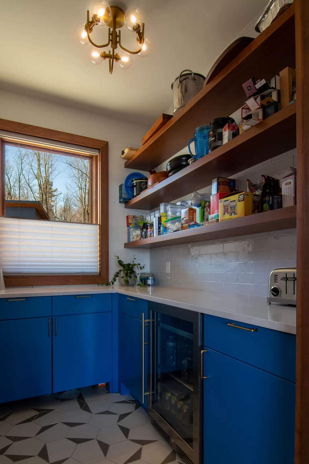 A modern pantry featuring bold blue cabinetry with gold hardware, white quartz countertops, and open wooden shelving stocked with food items, cookware, and kitchen accessories. A built-in beverage fridge is integrated into the lower cabinets. The backsplash consists of white subway tiles, and a stainless steel toaster sits on the counter. A large window with wood trim allows natural light to enter, partially covered by a pleated shade. The geometric-patterned floor complements the contemporary aesthetic, while a gold chandelier with exposed bulbs adds warmth and sophistication.