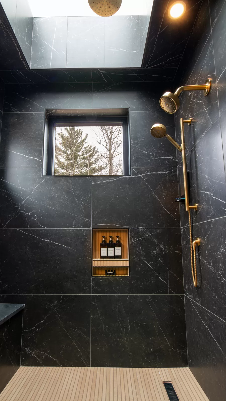 Luxury walk-in shower in Cinderwave, designed by AJA Architecture and Planning. This spa-inspired space features dramatic black marble walls with elegant white veining, contrasted by warm wood-textured flooring. A large skylight allows natural light to cascade into the shower, enhancing the open and airy ambiance. The gold shower system includes multiple showerheads, including a rainfall fixture, a handheld sprayer, and an adjustable wall-mounted option for a customized experience. A recessed wooden niche provides stylish and functional storage for bath essentials. The combination of dark stone, warm wood, and gold accents creates a bold yet serene retreat, blending modern sophistication with natural elements.