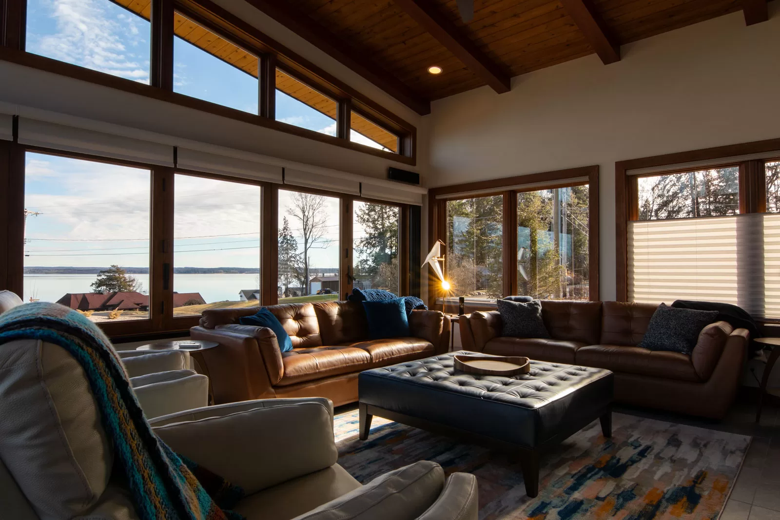 A modern living room with expansive floor-to-ceiling windows offering a scenic lake view. The space features warm wood accents, a vaulted ceiling with exposed beams, and natural light streaming in. Rich brown leather sofas with blue accent pillows create a cozy seating area around a large, tufted ottoman. A plush area rug with abstract patterns adds texture, while a sleek floor lamp provides ambient lighting. White armchairs with a striped knit throw enhance the inviting atmosphere. The panoramic windows frame the serene outdoor landscape, blending indoor comfort with the beauty of nature.